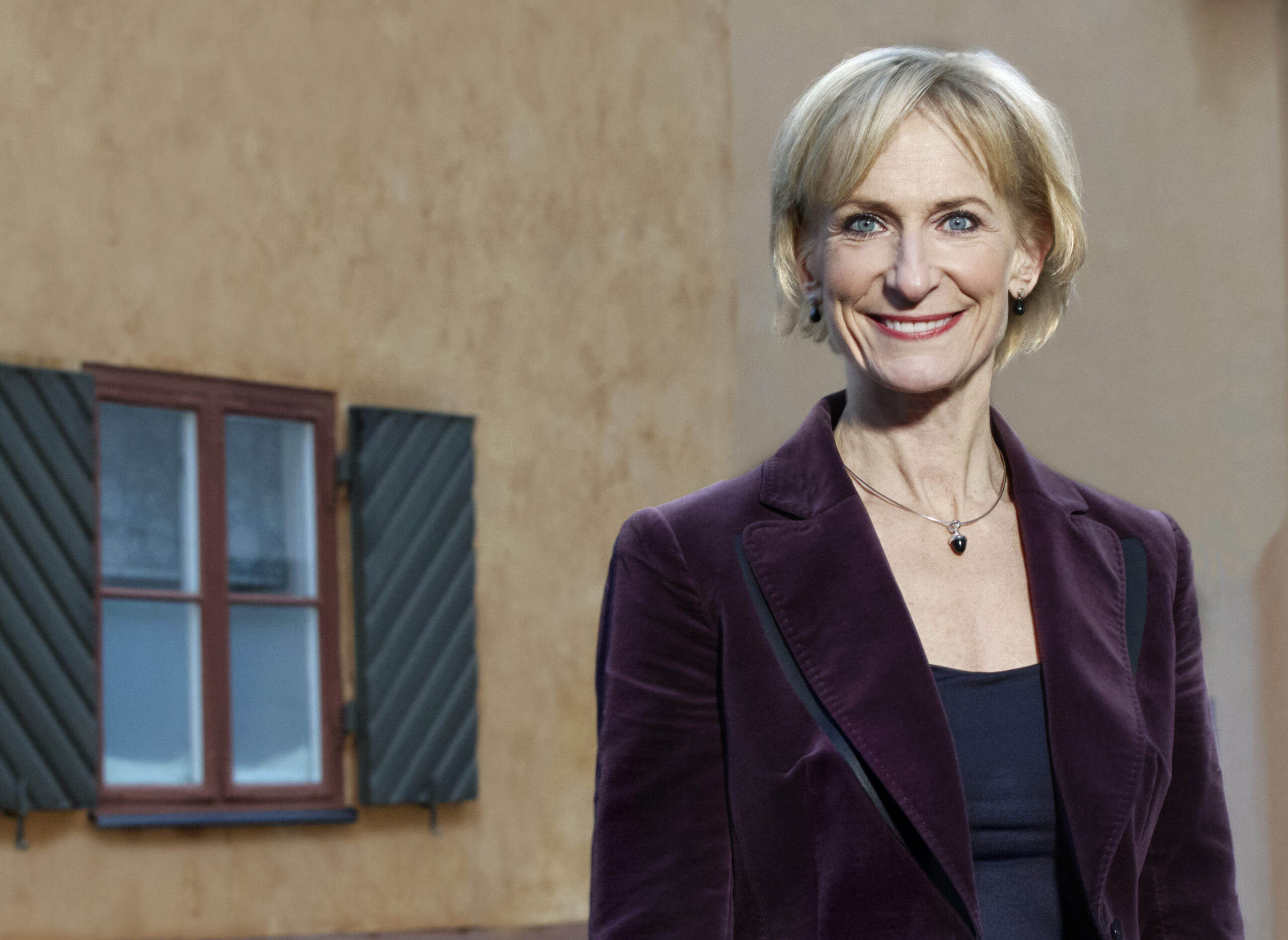 Kristina Alvendal, nationell industrisamordnare, ler mot kameran i mörklila sammetskavaj framför en gul husfasad med ett fönster med gröna fönsterluckor.