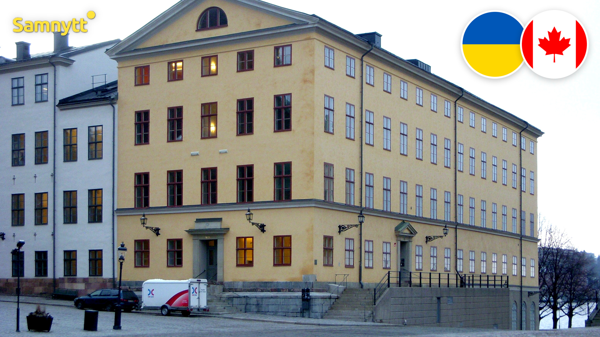 Gul stenbyggnad i flera våningar vid ett kullerstenstorg på Riddarholmen i Stockholm, med runda märken med Ukrainas och Kanadas flaggor uppe till höger.