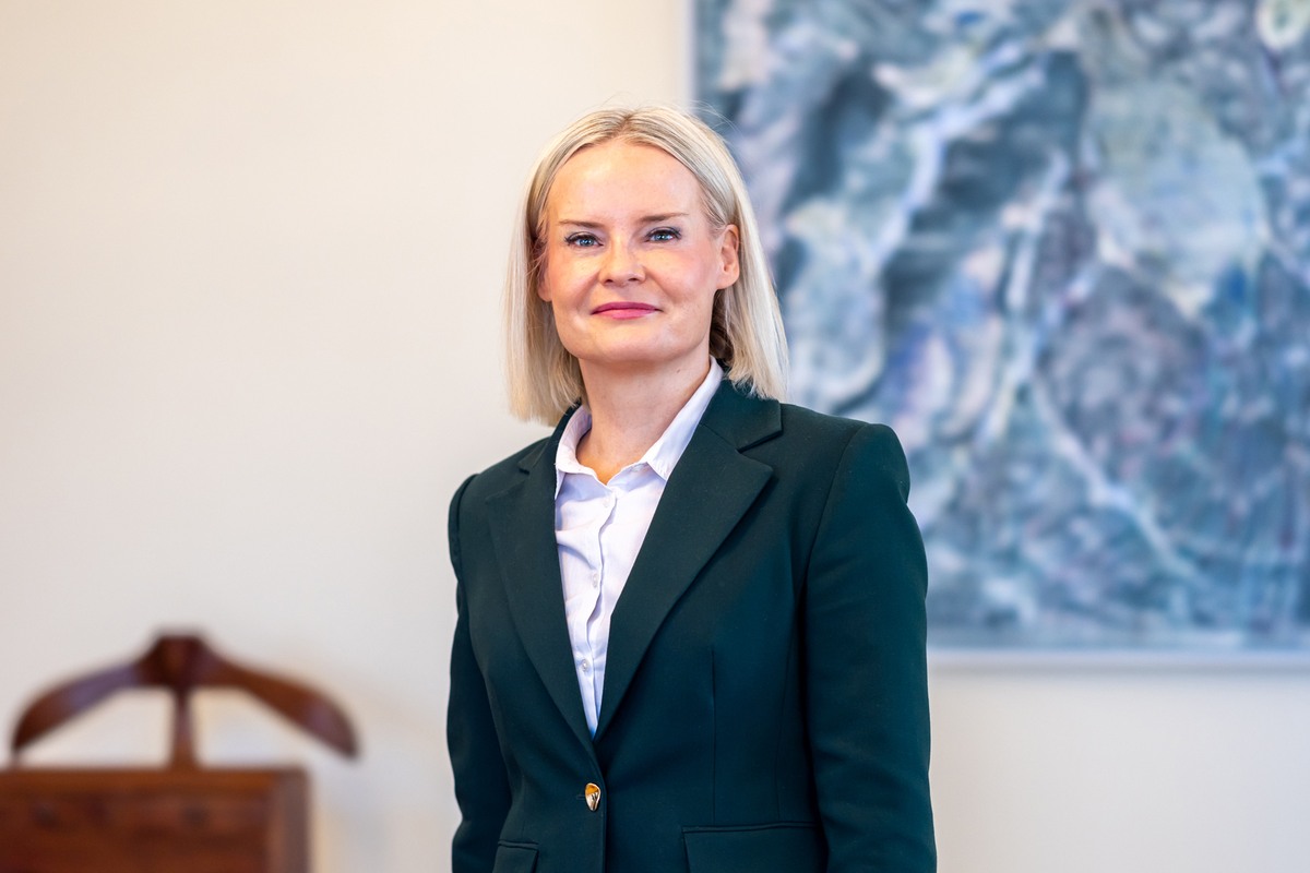 Riikka Purra i mörkgrön kavaj och vit skjorta, fotograferad i midjehöjd i ett ljust rum med en blå tavla i bakgrunden.